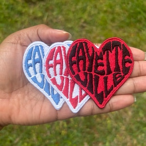 May include: Three heart-shaped patches with the word "FAYETTEVILLE" embroidered in a stylized font. One patch is white with blue text, another is white with red text, and the third is red with black text.