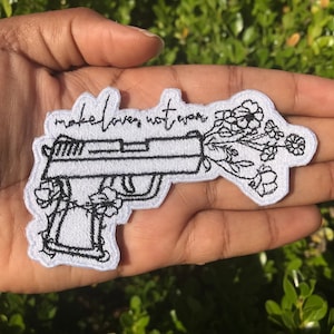 May include: White embroidered patch with a black outline of a gun with flowers growing out of the barrel. The text "make love not war" is written above the gun.