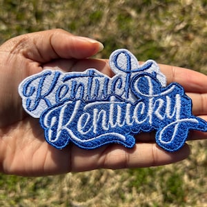 May include: A blue and white embroidered patch with the word "Kentucky" written in a cursive font.