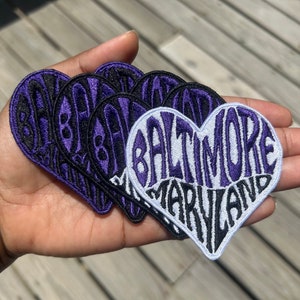 May include: A set of purple and white heart-shaped patches with the text "Baltimore Maryland" embroidered on them.