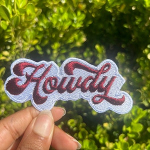 May include: A white embroidered patch with the word "Howdy" in red cursive lettering.