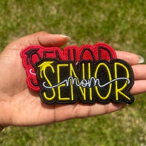 May include: Two iron-on patches with the text "Senior" in red and black with yellow outlines. The smaller patch has the text "Senior Mom" in white cursive writing.
