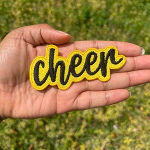 May include: A yellow felt patch with black embroidered lettering that reads "cheer".