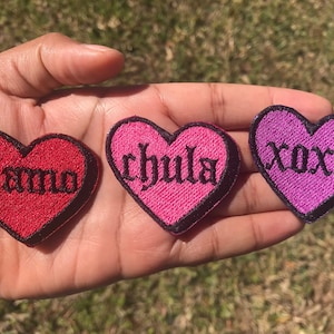 May include: Three heart-shaped embroidered patches with black gothic lettering. The patches are red, pink, and purple. The red patch says "te amo", the pink patch says "chula", and the purple patch says "xoxo".