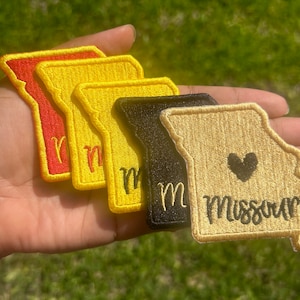 May include: Four embroidered patches in the shape of the state of Missouri. The patches are in red, yellow, black, and beige. The patches have the word "Missouri" and a heart embroidered on them.