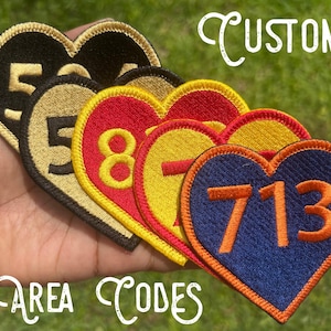 May include: Heart-shaped embroidered patches with area codes. The patches feature various color combinations, including black and gold, red and yellow, and blue and orange. The word "CUSTOM" and "AREA CODES" are written in white script.