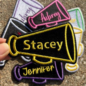May include: Embroidered megaphone patches in various colors, each with a name in a contrasting color. The patches are black, purple, pink, and white, with names like "Stacey" and "Aubrey" in yellow and pink. The patches are likely for cheerleading or similar activities.