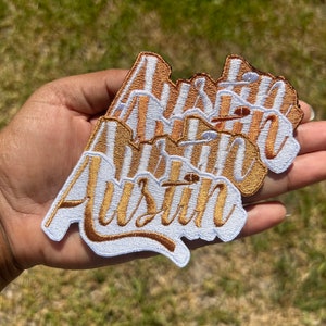 May include: Three embroidered patches featuring the word Austin in a cursive script. The lettering is rendered in a metallic gold thread with a white border and a small football icon integrated into the design above the letter i.