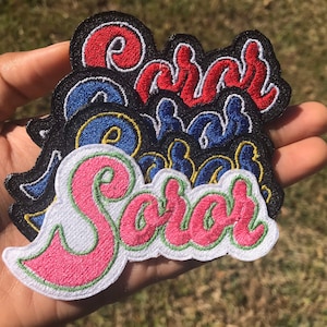 May include: A set of five embroidered iron-on patches with the name "Sarah" in different colors and styles. The patches are in a hand, with the pink and green patch in the foreground.