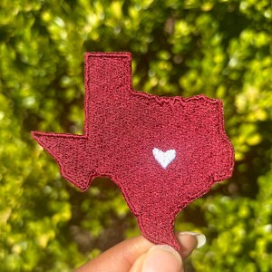 May include: A maroon embroidered patch in the shape of the state of Texas, featuring a white heart in the center. The patch is held against a blurred green background.