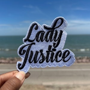 May include: A white embroidered patch with black lettering that reads "Lady Justice".