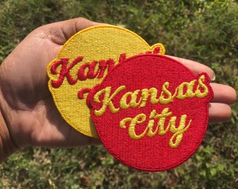 Puede incluir: Dos parches bordados, uno amarillo con letras rojas que dicen "Kansas" y uno rojo con letras doradas que dicen "Kansas City".