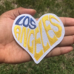 Peut inclure: Un patch en forme de cœur blanc avec les mots "LOS ANGELES" brodés en jaune et bleu.