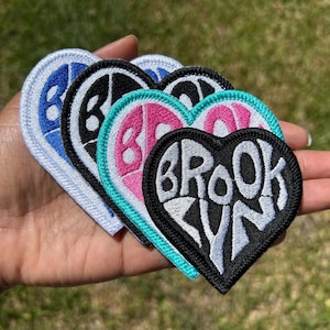 May include: A collection of four heart-shaped embroidered patches featuring the word BROOKLYN in stylized lettering. The patches come in various color combinations including blue and white, black and white, pink and teal, and black and white.