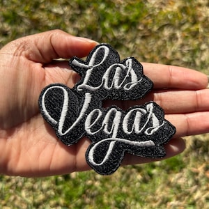 May include: Black and white embroidered patch with the text "Las Vegas" in a cursive font.