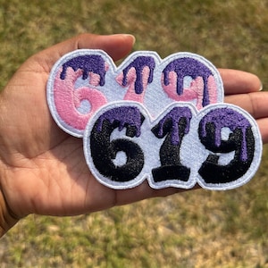 Peut inclure: Trois patchs thermocollants avec les chiffres 6, 7 et 9. Les chiffres sont noirs avec un contour violet et de la peinture violette qui coule. Les patchs sont sur un fond blanc.