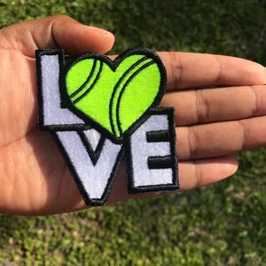 Peut inclure: Patch en feutre blanc avec le mot "LOVE" en lettres noires soulignées. Une balle de tennis verte en forme de cœur, chevauchant les lettres.