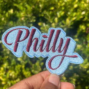 May include: A blue and maroon embroidered patch with the word "Philly" in a cursive font.