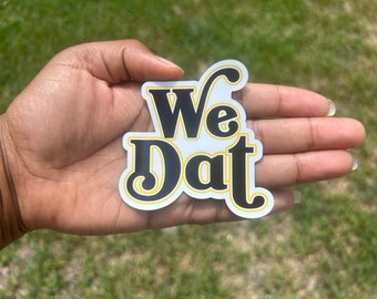 May include: A white sticker with black text that reads "We Dat" in a retro font with a yellow outline.