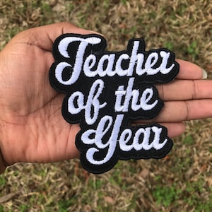 May include: White embroidered text on a black patch that reads "Teacher of the Year".