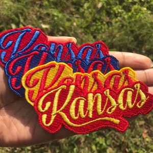 May include: Three embroidered patches with the word "Kansas" in a stylized font. The patches are layered on top of each other, with the top patch being red, the middle patch being yellow, and the bottom patch being blue.