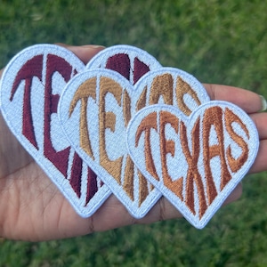 May include: Four heart-shaped embroidered patches with the word "TEXAS" in various colors. The patches have a white border and are held in a hand against a green background.