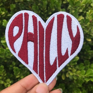 May include: A white heart-shaped patch with the word "PHILLY" embroidered in maroon.