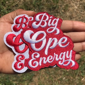 May include: Embroidered patches with the words "Big Cope Energy" in white script on a red background. The patches have a white border and are held in a hand against a blurred green background.