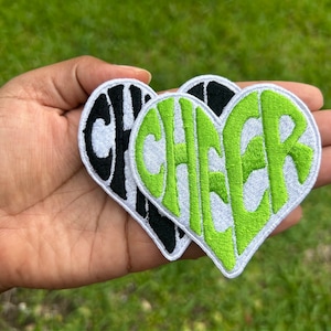 May include: Two heart-shaped patches with the word "CHEER" embroidered in a white outline. One patch is black with white lettering, and the other is white with green lettering.