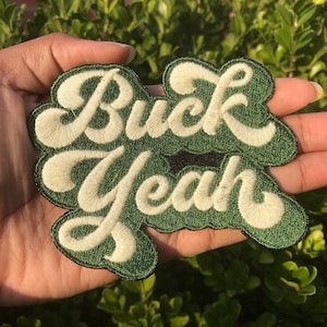 May include: Green embroidered patch with white lettering that reads "Buck Yeah" in a retro font.