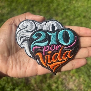 May include: Two heart-shaped embroidered patches. One is gray and white, the other is black with teal, pink, and orange text that reads "210 por Vida". The patches are held in a hand against a green background.