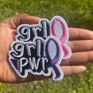 May include: Embroidered patches with the words "grl PWR" in white, black, and purple, with a pink and purple ribbon design. The patches have a white border. The patches are held in a hand.