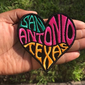 May include: Embroidered heart-shaped patch with the words "SAN ANTONIO TEXAS" in teal, pink, and yellow stitching on a black background. The patch is held in a hand against a blurred green background.