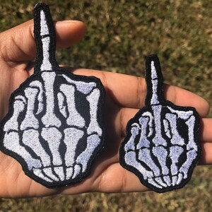 May include: Two embroidered iron-on patches of a skeleton middle finger, one larger than the other. Both patches are white on black fabric.