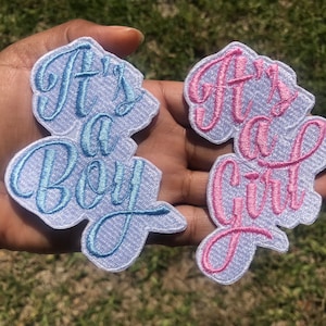 May include: Two white fabric patches with blue and pink embroidery. The left patch says "It's a Boy" in blue embroidery. The right patch says "It's a Girl" in pink embroidery.