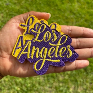May include: Two embroidered patches with the words "Los Angeles" in yellow thread on a purple background.