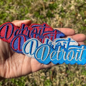 May include: Embroidered patches in red, white, and blue, spelling "Detroit" in a cursive font. The patches are in a variety of colors, including red, blue, and white. The patches are likely for clothing or accessories.