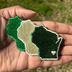 May include: Three embroidered patches in the shape of the state of Wisconsin. The patches are green, white, and a darker green. The darker green patch has a small white heart in the center.