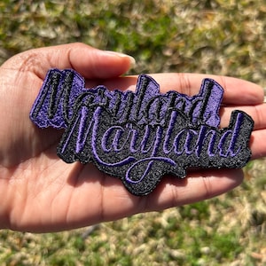 May include: Purple and black embroidered patch with the word "Maryland" written twice in a stylized font.