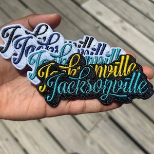 Peut inclure: Un ensemble de cinq patchs brodés avec le mot "Jacksonville" dans différentes couleurs et styles. Les patchs sont empilés les uns sur les autres, le plus grand patch en bas et le plus petit patch en haut. Tous les patchs sont noirs avec des broderies de différentes couleurs.