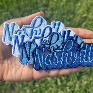 May include: A set of five embroidered patches in shades of blue, each featuring the word "Nashville" in a cursive script. The patches are layered on top of each other, with the lightest blue patch on top and the darkest blue patch on the bottom.