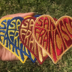 May include: Four heart-shaped embroidered patches with the text "San Francisco" in a stylized font. The patches are in yellow, blue, red, and gold.