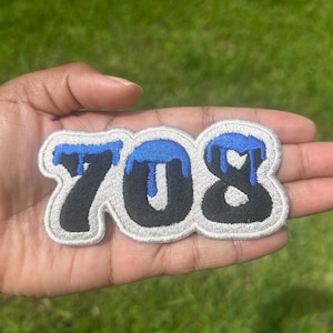 May include: An embroidered patch featuring the numbers 708 in black with a silver-grey border. The top portions of the numbers are accented with a bright blue dripping paint effect, creating a distinct visual contrast against the dark numerals.