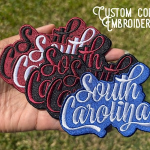 May include: Embroidered patches in red, black, and blue, each featuring the words "South Carolina" in a cursive script. The patches have a white border and are held in a hand. The text "CUSTOM COLOR EMBROIDERY" is in the background.