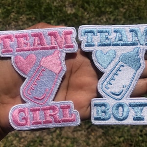 May include: Two white iron-on patches with blue and pink embroidery. The left patch says "Team Girl" with a pink heart and a baby bottle. The right patch says "Team Boy" with a blue heart and a baby bottle.