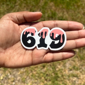 May include: A white sticker with the number "619" in black, with pink dripping accents. The sticker is held in a hand against a background of green grass.