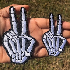 May include: Two white skeleton hand peace sign patches. The larger patch is about 4 inches tall and the smaller patch is about 2 inches tall.