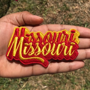 May include: Embroidered patch with the word "Missouri" in a decorative script. The patch has a red base with yellow lettering and a yellow border. The patch is held in a hand.