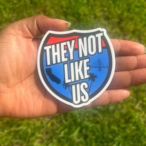 May include: A white sticker with a blue and red design. The sticker is shaped like a highway sign and says "They Not Like Us" with a silhouette of California and the Golden Gate Bridge.