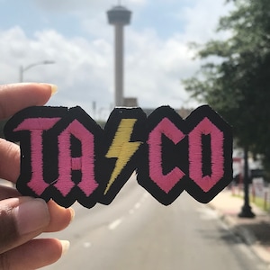 May include: A black and pink embroidered patch with the word "TACO" in pink letters and a yellow lightning bolt in the middle. The patch is held in a hand with a blurred background.
